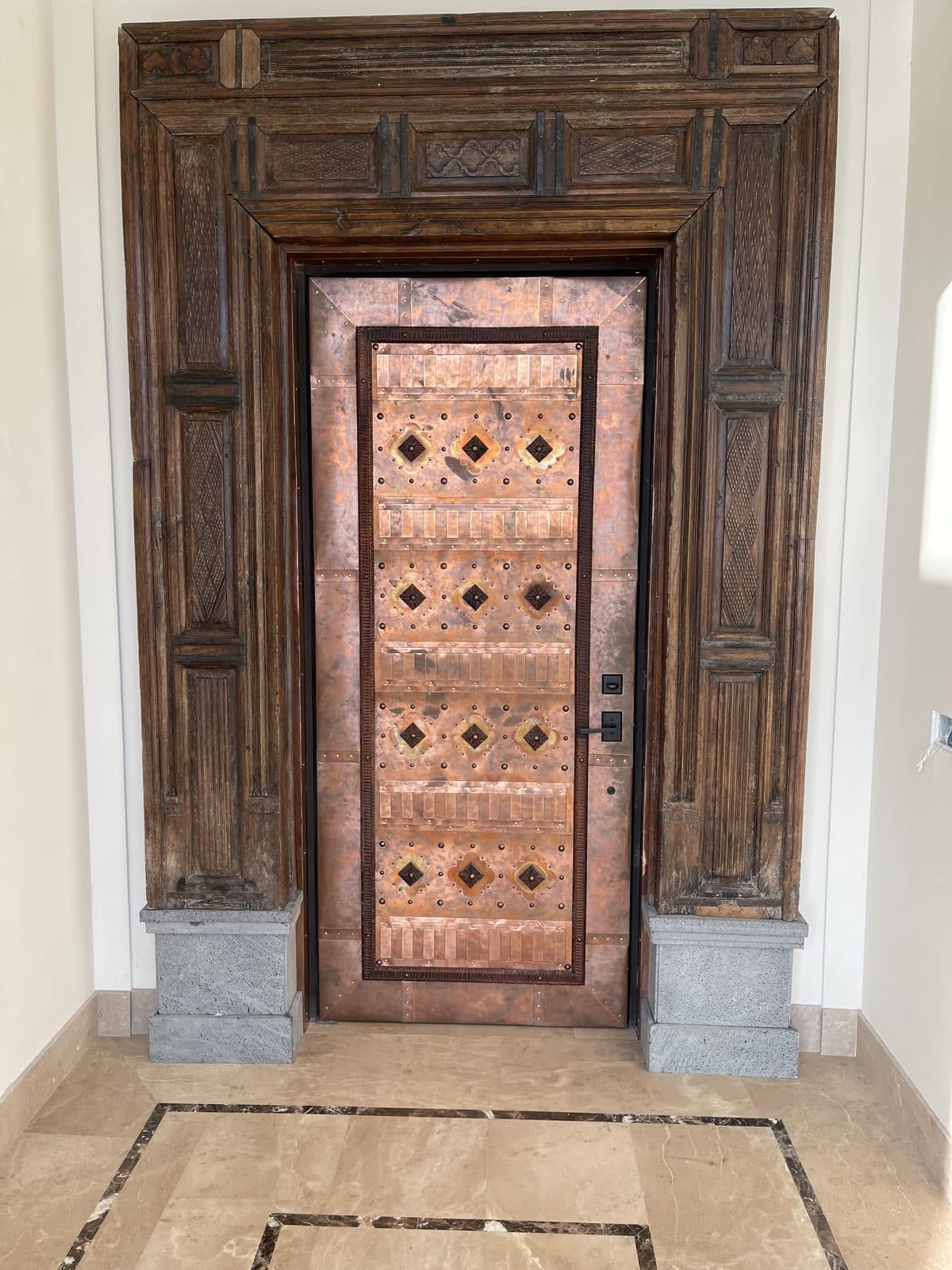 Copper front door with geometric bronze inlays, framed by an antique hand-carved wood surround from India
