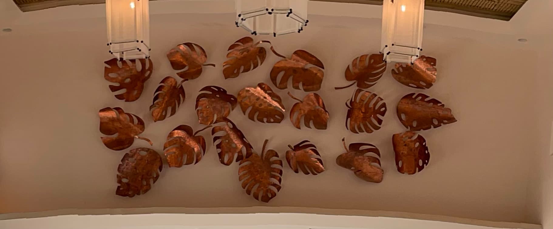 Copper mano de tigre wall installation above the onyx dining table at Villa Luciérnaga -- about twenty hand-fabricated copper leaves arranged on the wall where the great room's soaring ceiling steps down toward the kitchen